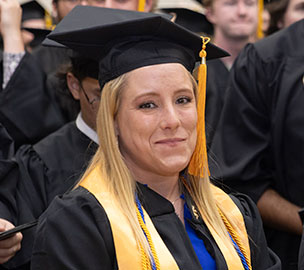Graduate wearing a black cap and gown with a yellow honor cord smiles during a commencement ceremony.