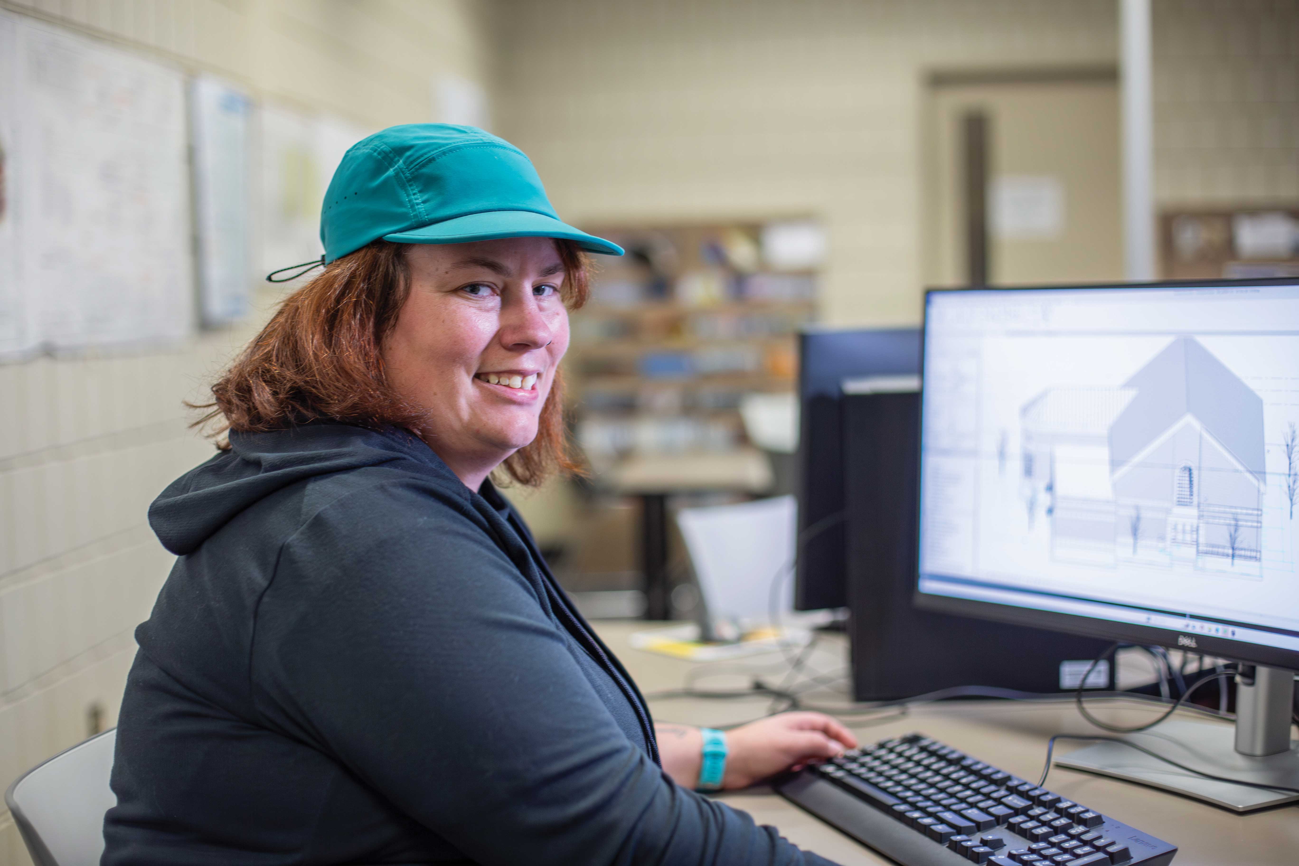 Person wearing a green cap sitting at a desk with a computer displaying architectural design software.